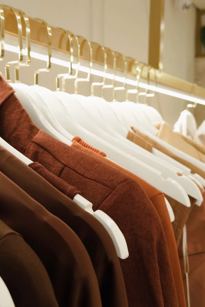 Detalhe dos cabides brancos com blazers e camisas em marrom, telha e caramelo