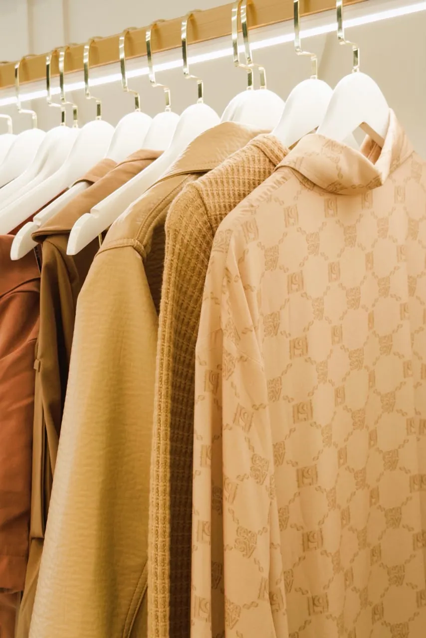 Detalhe de jaquetas e camisas camel com monograma alinhadas em cabides brancos